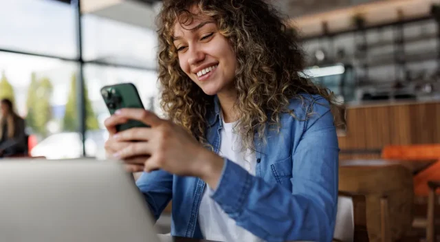 Chica-joven-sonriente-utilizando-su-movil-mientras-trabaja-con-su-portatil-en-una-cafeteria-Young-smiling-woman-using-mobile-phone-while-working-on-a-laptop-at-a-cafe